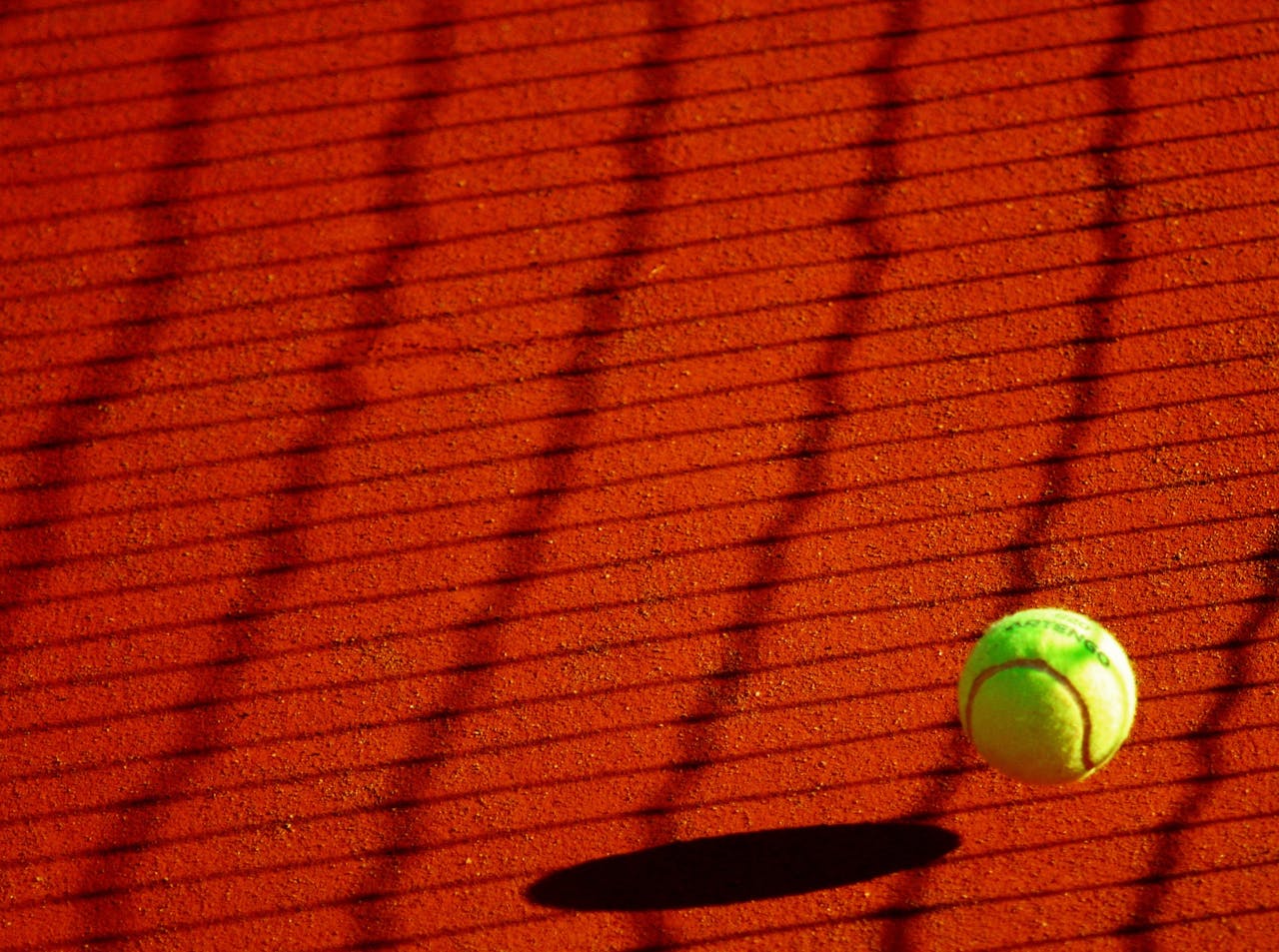 Home A vibrant tennis ball casting a shadow on a sunlit clay court with net shadows.